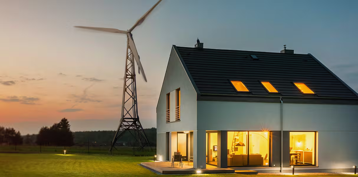 Haus mit installierter kleinen Windturbine und Solarpaneelen bei Sonnenuntergang – Beispiel für Mikronetz mit Windenergie.