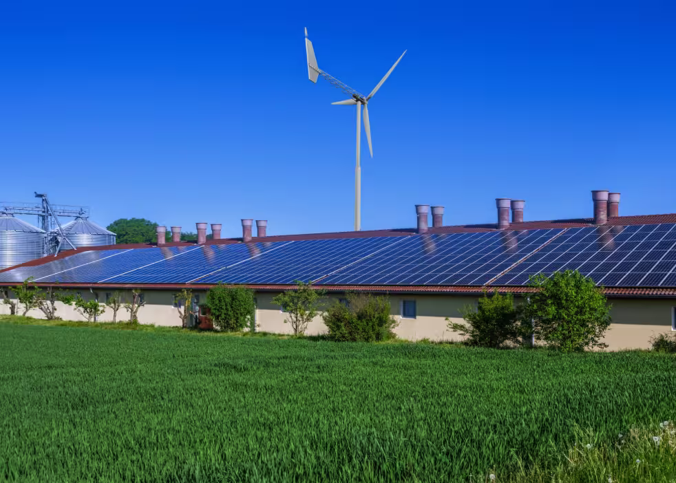 Modernes landwirtschaftliches Gebäude mit Solarmodulen auf dem Dach und einer vertikalen Windturbine daneben – ein hybrides System für erneuerbare Energien im ländlichen Raum.
